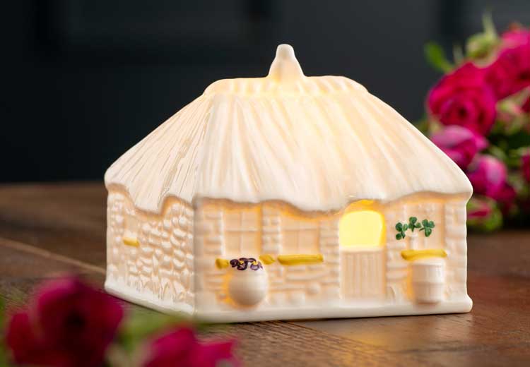 A ceramic Irish cottage, illuminated from the inside, decorated with painted shamrocks and flowers on a table with roses