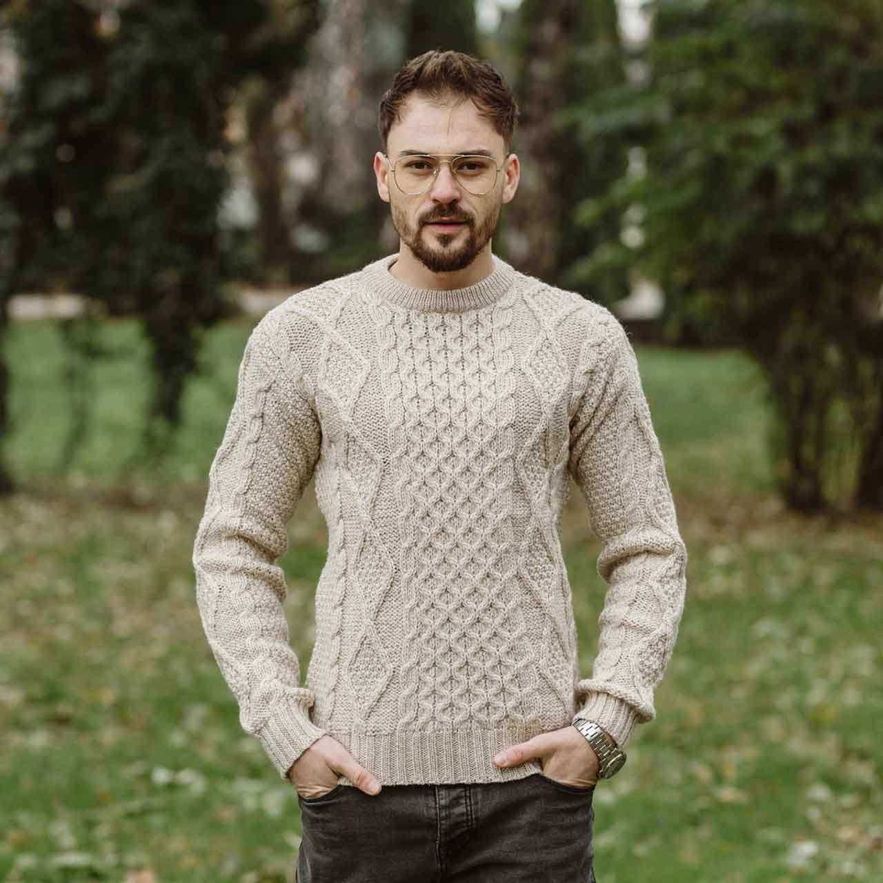 man wearing a parsnip colored fisherman cable knit mens sweaterstanding outdoors with trees in the background