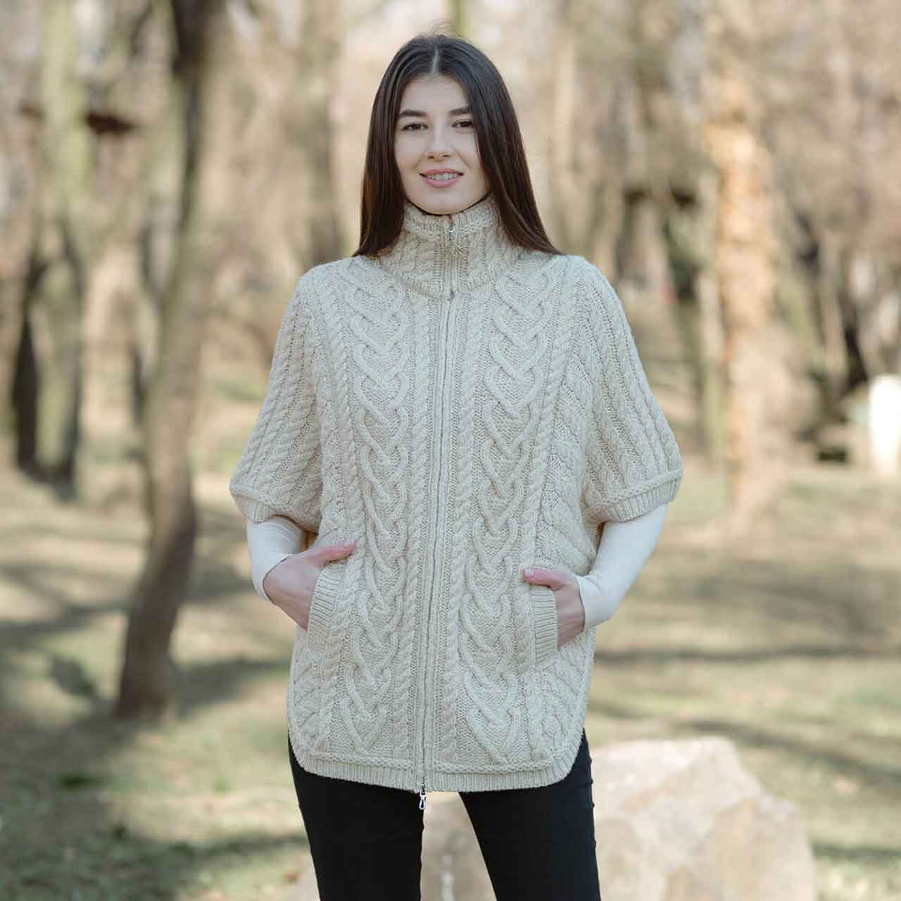 woman wearing an aran knit funnel neck poncho jacket, natural-colored, in a forest setting