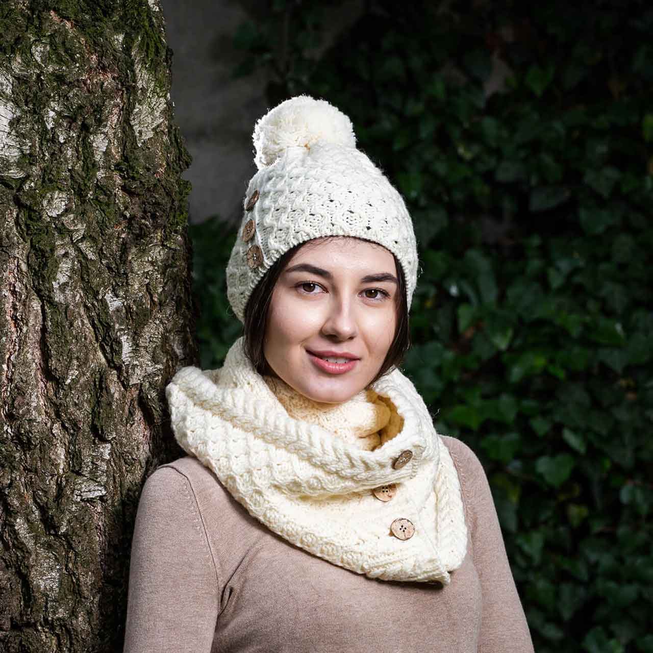 woman wearing a natural colored merino anan knit button pom-pom hat and matching scarf, leaning against a tree