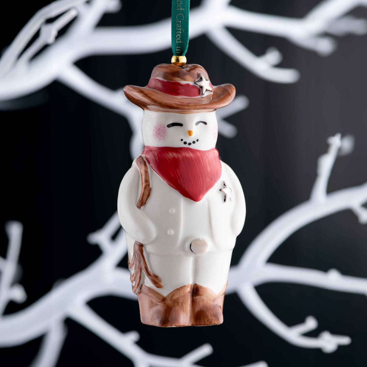 belleek wild west snowman ornament a decorative snowman ornament with a cowboy hat and red scarf against a dark background with white branches.