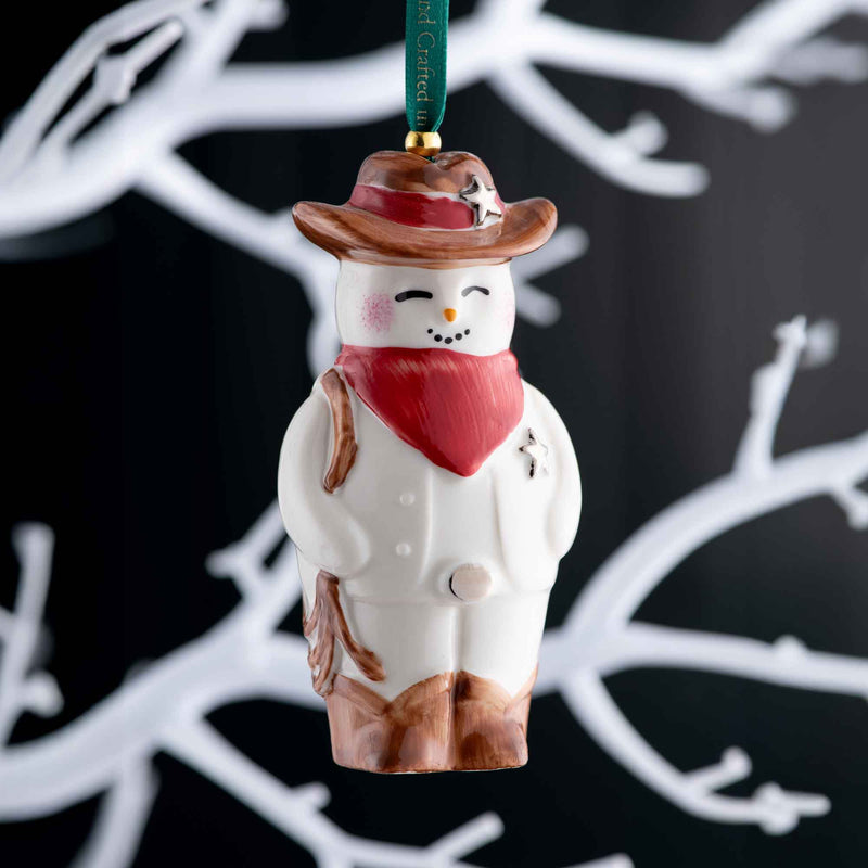 Belleek Wild West Snowman Ornament A Decorative snowman ornament with a cowboy hat and red scarf against a dark background with white branches.
