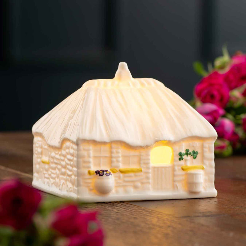 Belleek 2025 LED Irish Thatched Cottage A Cottage-shaped night light on a wooden surface with flowers in the background