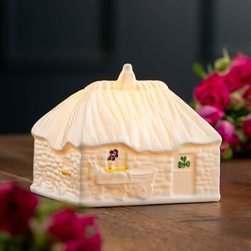 2025 LED Irish Thatched Cottage A Cottage-shaped night light with shamrock design, with details like a donkey and cart on a wooden surface with flowers.
