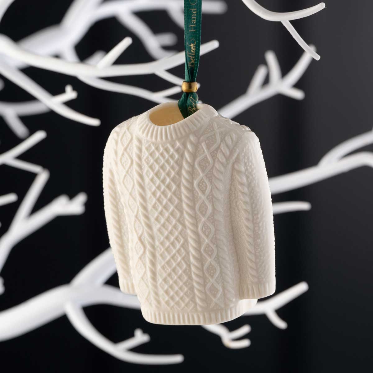 belleek pottery aran jumper christmas ornament, on a green ribbon, hanging on a white tree against a black background.
