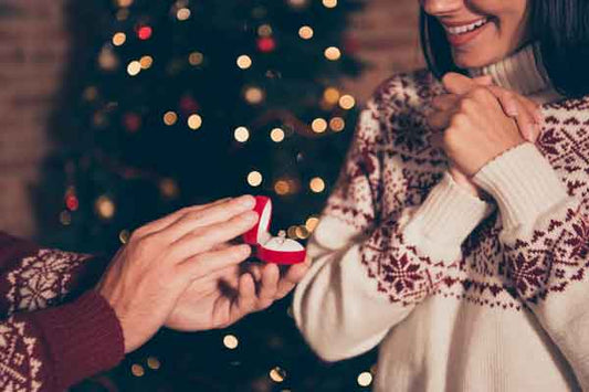 Close up of a man proposing to a woman in front of a Christmas tree. Both are wearing Christmas sweaters
