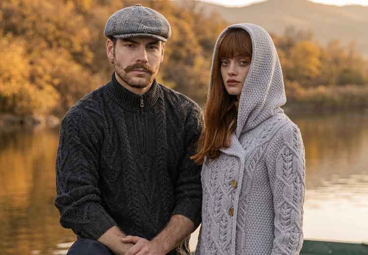 A man and a woman wearing Irish Knitwear sitting together outdoors by a lake at sunset. The man wears a dark Irish Aran Knit Fisherman sweater and a flat cap, while the woman wears a gray, hooded Irish Aran Leaf Coat
