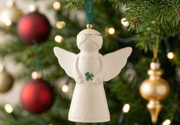 A white ceramic Belleek Pottery Angel Hanging Ornament hanging from a green tree branch. The ornament features a small green shamrock on the chest and a green hanging ribbon.