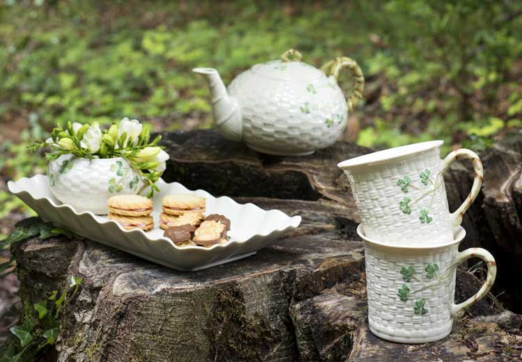 Belleek Pottery teapot, mugs and tray painted with signature shamrock, on a stone surface with greenery in the background.