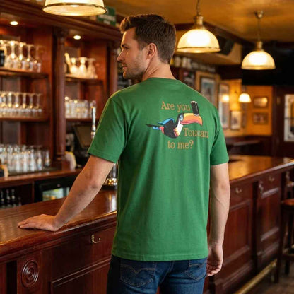 Man standing at a bar with his back to the camera wearing a Guinness® Are you Toucan to Me Green T-shirt with a toucan graphic and 'Are you Toucan to me?' text