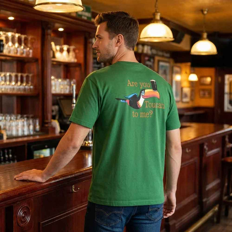 Man standing at a bar with his back to the camera wearing a Guinness® Are you Toucan to Me Green T-shirt with a toucan graphic and 'Are you Toucan to me?' text