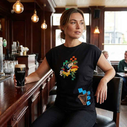 Woman sitting at a bar holding a Guinness glass, wearing a black Guinness® Hawaiian Ruched Ladies Black T-shirt