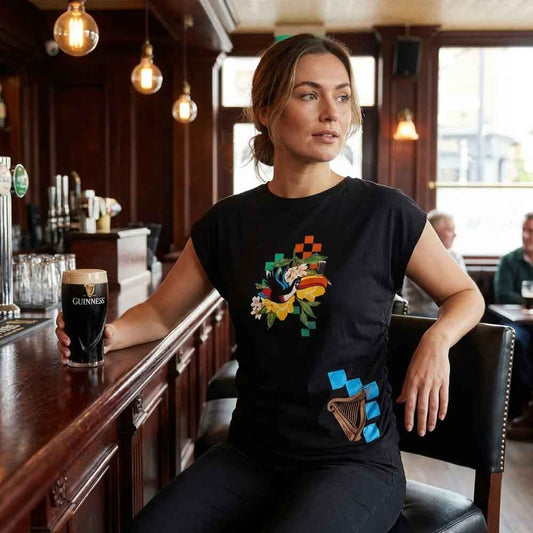 Woman sitting at a bar holding a Guinness glass, wearing a black Guinness® Hawaiian Ruched Ladies Black T-shirt