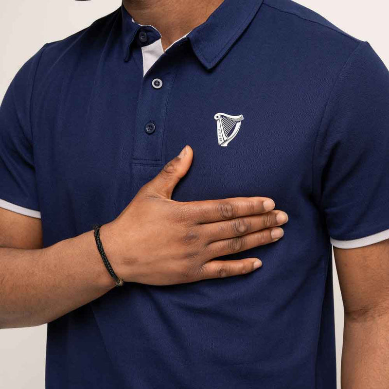 Person wearing a Guinness® Navy Performance Polo Shirt, with their hand beneath the Harp Logo on the left hand side of the chest