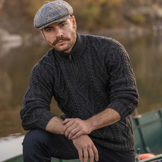 A man wearing a charcoal Merino Wool Aran Knit Zip Neck Fisherman Mens Sweater and a patterned flat cap, sitting outdoors on a boat with a blurred natural background.