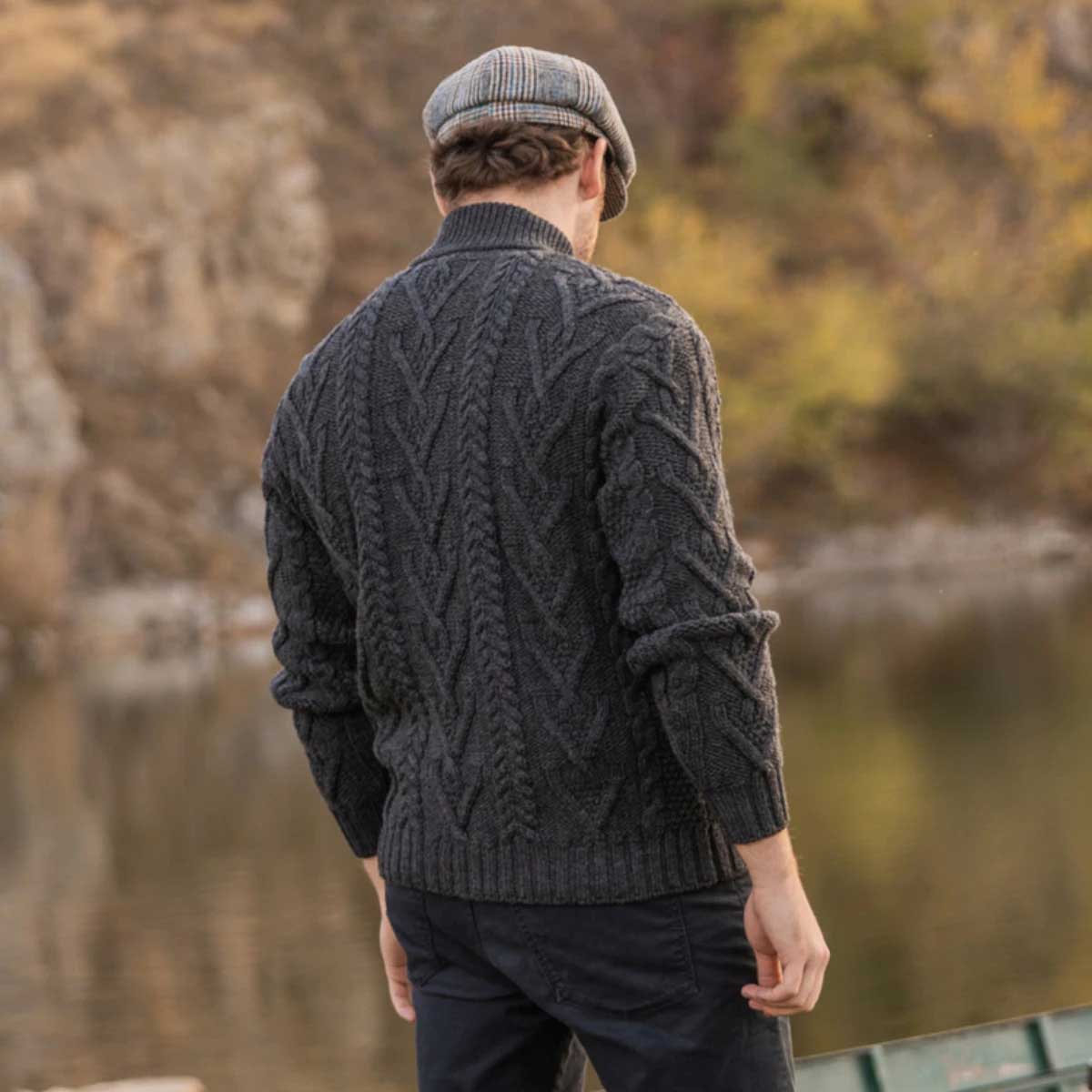 a man viewed from behind wearing a charcoal merino wool aran knit zip neck fisherman men's sweater and a gray flat cap, standing outdoors near a body of water with a blurred natural background.