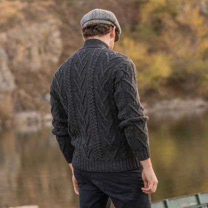 A man viewed from behind wearing a charcoal Merino Wool Aran Knit Zip Neck Fisherman Men's Sweater and a gray flat cap, standing outdoors near a body of water with a blurred natural background.