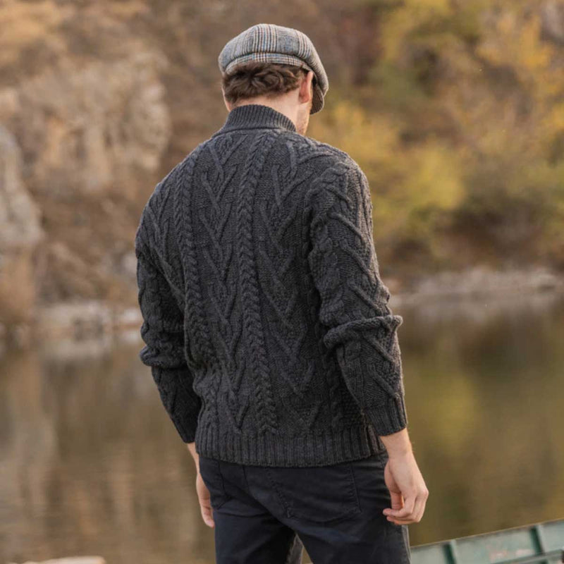 A man viewed from behind wearing a charcoal Merino Wool Aran Knit Zip Neck Fisherman Men's Sweater and a gray flat cap, standing outdoors near a body of water with a blurred natural background.