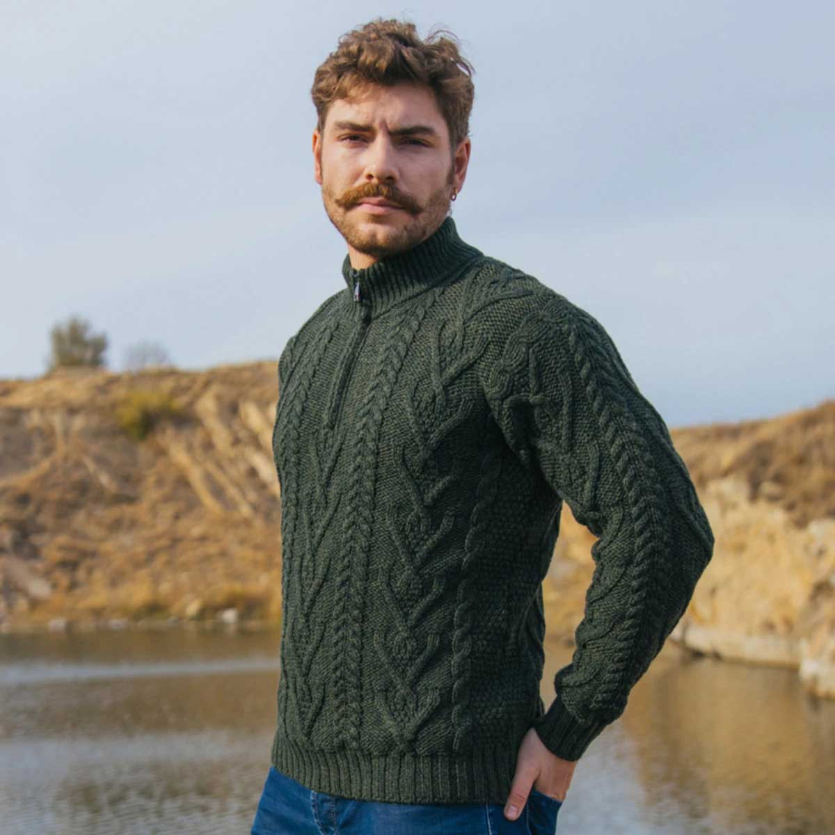 a man wearing a merino wool aran knit zip neck fisherman mens sweater in army green, standing outdoors in front of a dry, sandy hillside and water.