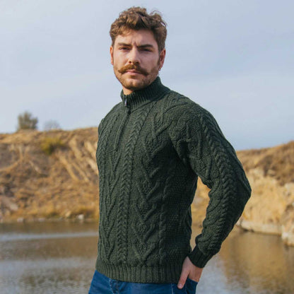 A man wearing a Merino Wool Aran Knit Zip Neck Fisherman Mens Sweater in Army Green, standing outdoors in front of a dry, sandy hillside and water.
