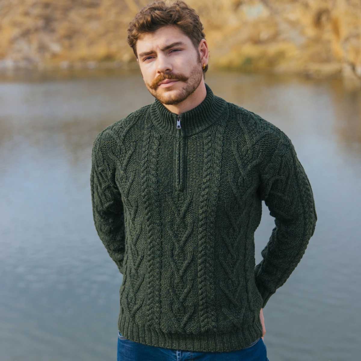 a man wearing an army green merino wool aran knit zip neck fisherman mens sweater, standing outdoors with a lake and rocky shoreline in the background.
