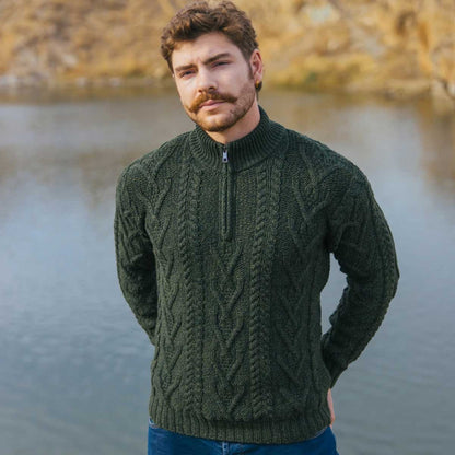 A man wearing an Army Green Merino Wool Aran Knit Zip Neck Fisherman Mens Sweater, standing outdoors with a lake and rocky shoreline in the background.