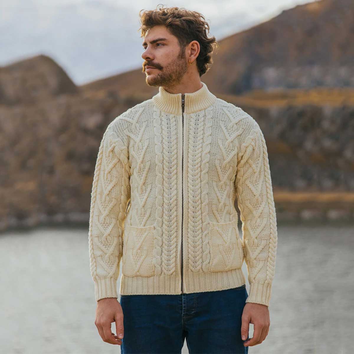 a man wearing a natural color merino wool aran knit mens zip cardigan and front pockets, standing outdoors in front of a blurred mountain and water background.