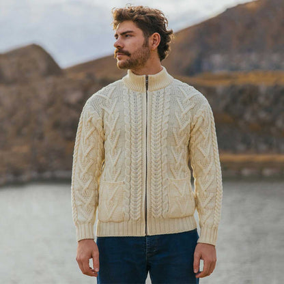 A man wearing a natural color Merino Wool Aran Knit Mens Zip Cardigan and front pockets, standing outdoors in front of a blurred mountain and water background.