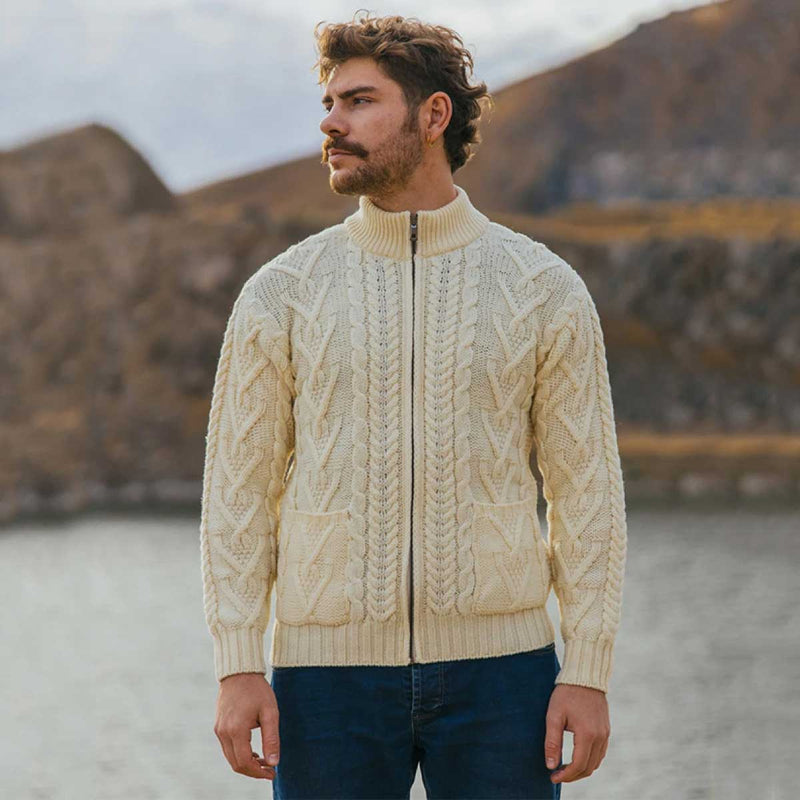A man wearing a natural color Merino Wool Aran Knit Mens Zip Cardigan and front pockets, standing outdoors in front of a blurred mountain and water background.