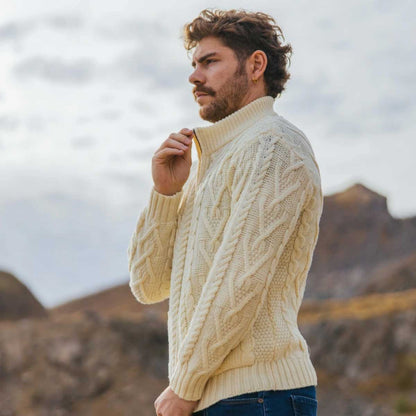 A man wearing a natural-colored Merino Wool Aran Knit Mens Zip Cardigan, standing outdoors against a soft-focus rocky landscape background.