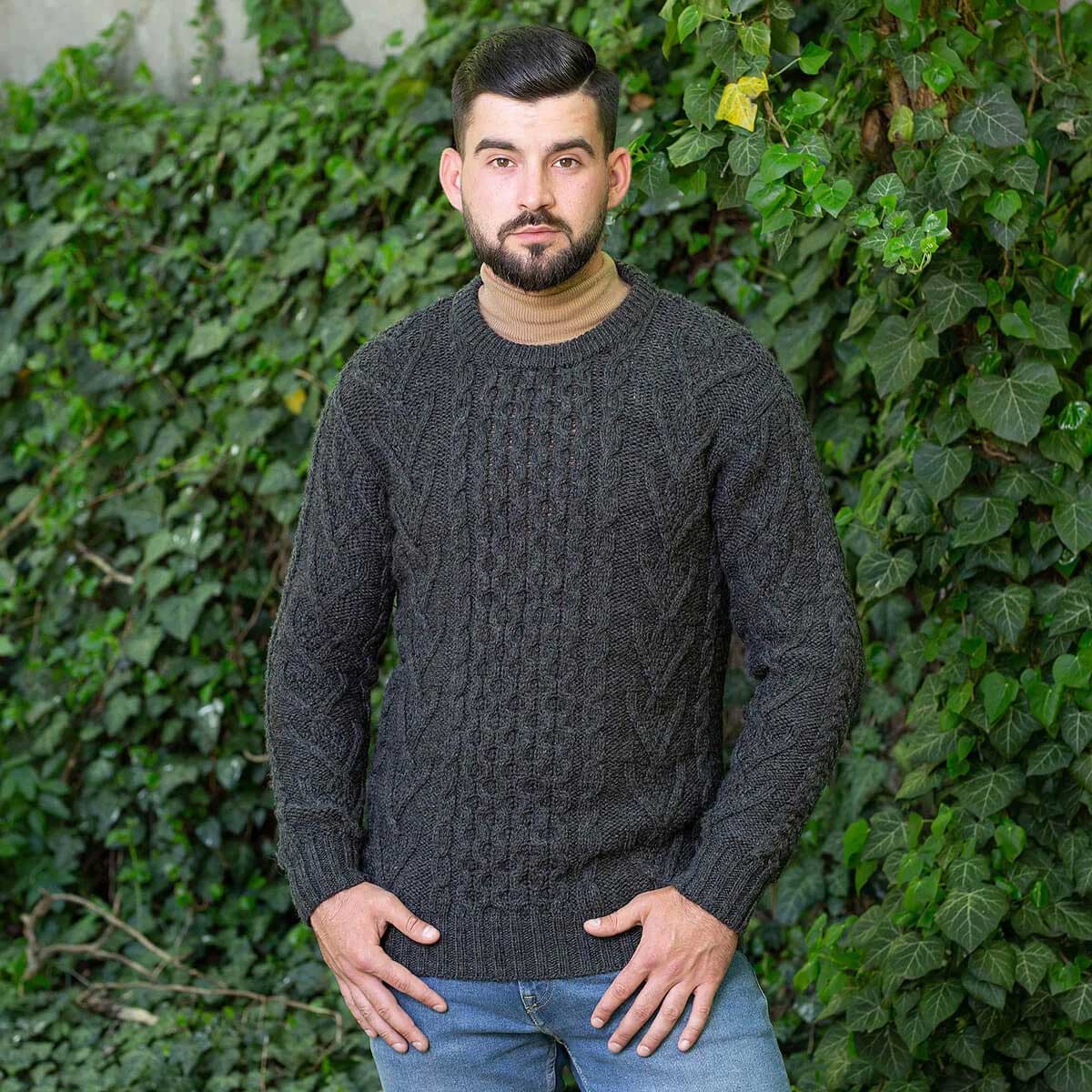 a man wearing a charcoal merino wool classic aran knit men's sweater standing in front of a green ivy-covered wall.