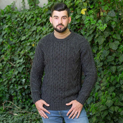 A man wearing a charcoal Merino Wool Classic Aran Knit Men's Sweater standing in front of a green ivy-covered wall.