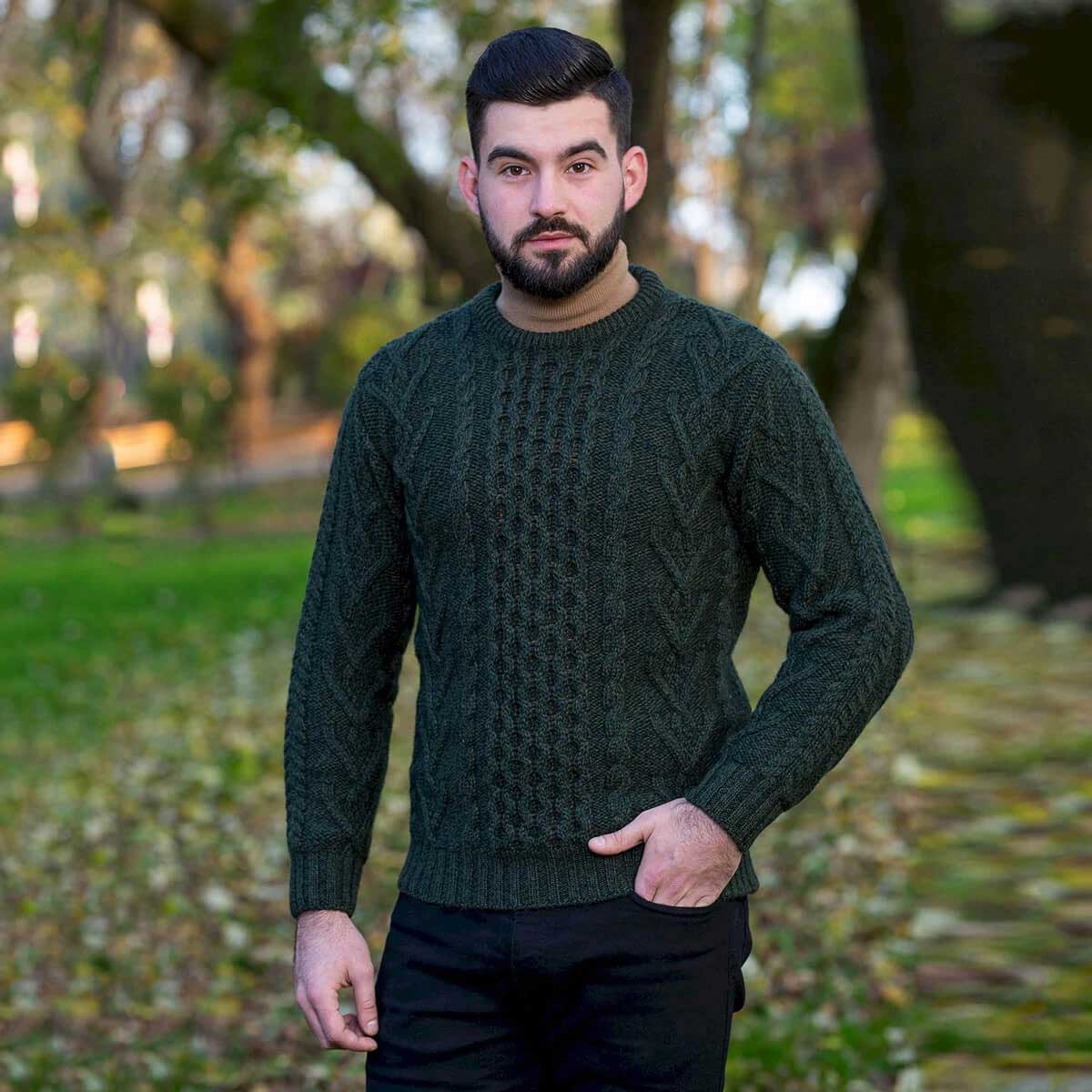 a man wearing an army green cable-knit merino wool classic aran knit men's sweater standing in a blurred park setting with trees and fallen leaves.