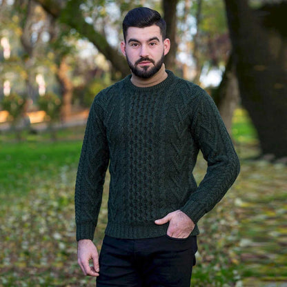 A man wearing an Army Green cable-knit Merino Wool Classic Aran Knit Men's Sweater standing in a blurred park setting with trees and fallen leaves.