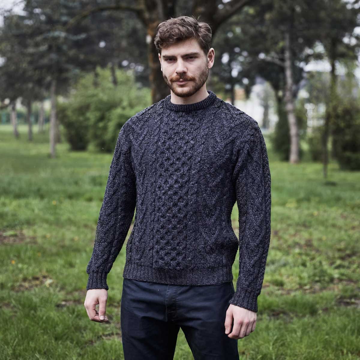 a man wearing a charcoal-colored merino wool classic aran knit men's sweater standing in a park