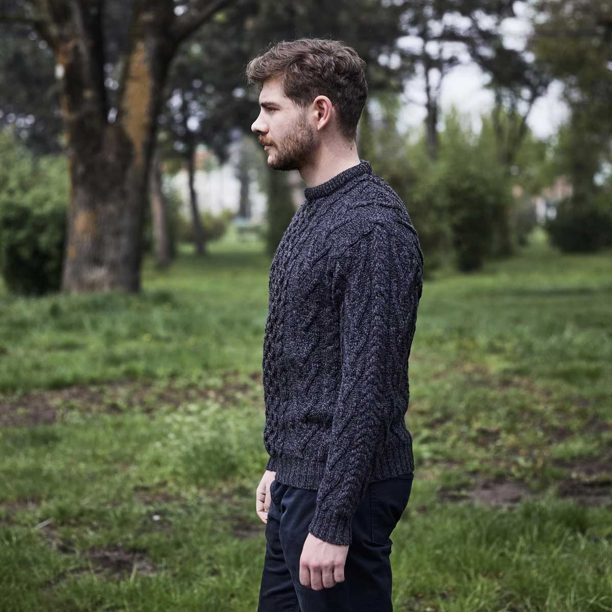 a man wearing a charcoal-colored merino wool classic aran knit men's sweater stands in profile in a grassy park with trees in the background.