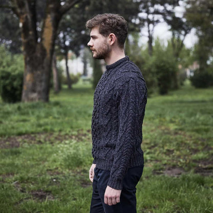 A man wearing a charcoal-colored Merino Wool Classic Aran Knit Men's Sweater stands in profile in a grassy park with trees in the background.