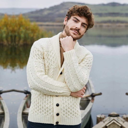 A man wearing a natural-colored Aran Cable Knit Shawl Collar Mens Cardigan, standing by a calm lake with boats and hills in the background.