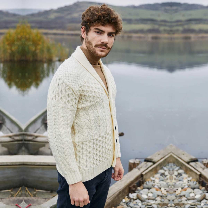 A man wearing a natural-colored Aran Cable Knit Shawl Collar Mens Cardigan over a tan shirt and dark trousers, standing outdoors in front of a calm lake with boats and hills in the background.