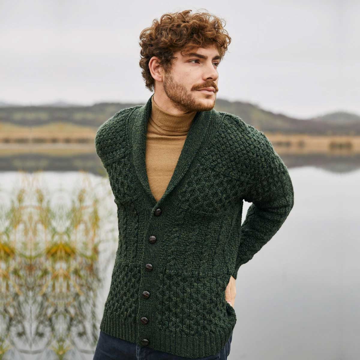 a man wearing an army green aran cable knit shawl collar mens cardigan over a tan turtleneck, standing outdoors near a calm lake with hills in the background.