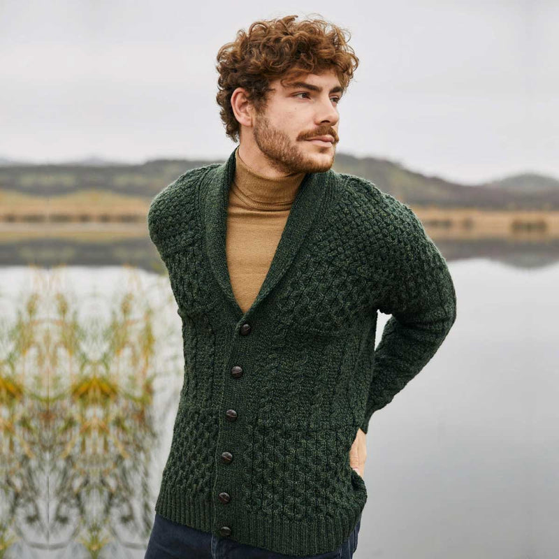 A man wearing an Army Green Aran Cable Knit Shawl Collar Mens Cardigan over a tan turtleneck, standing outdoors near a calm lake with hills in the background.