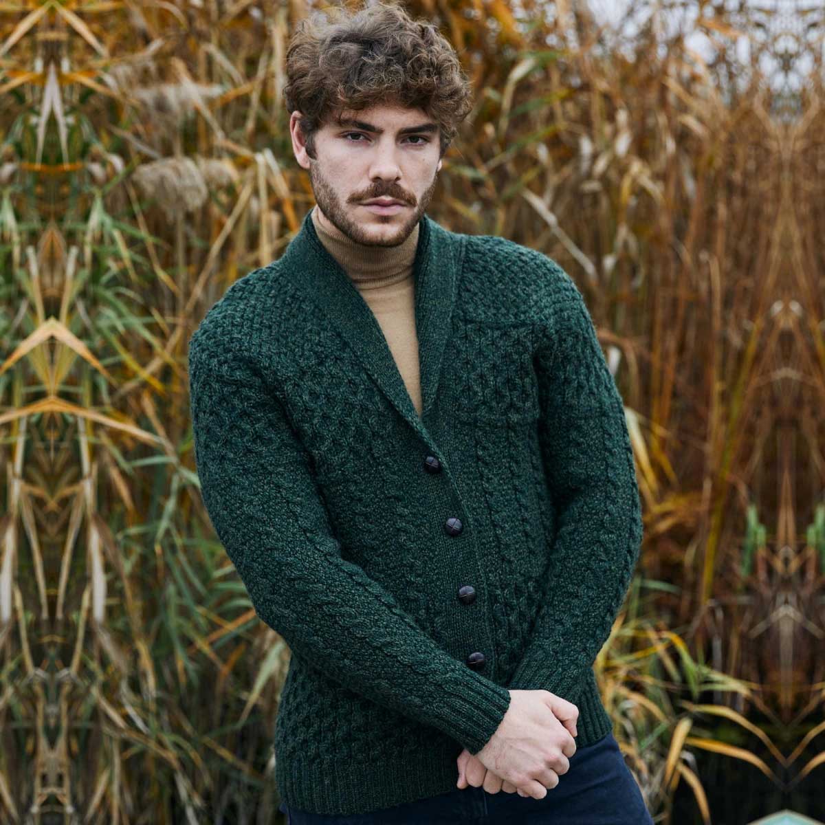 a man wearing an army green aran cable knit shawl collar mens cardigan over a tan turtleneck, standing outdoors with tall grass or reeds in the background