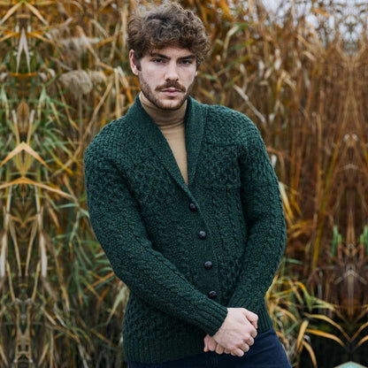 A man wearing an Army Green Aran Cable Knit Shawl Collar Mens Cardigan over a tan turtleneck, standing outdoors with tall grass or reeds in the background