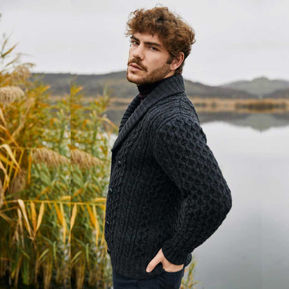 A man wearing a charcoal Double Breasted Shawl Collar Aran Cable Knit Mens Cardigan stands in profile near a calm lake with reeds in the background.