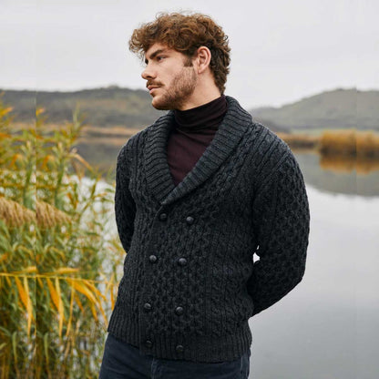 A man wearing a charcoal Double Breasted Shawl Collar Aran Cable Knit Mens Cardigan over a dark turtleneck, standing outdoors near a calm lake with reeds and distant hills under an overcast sky.