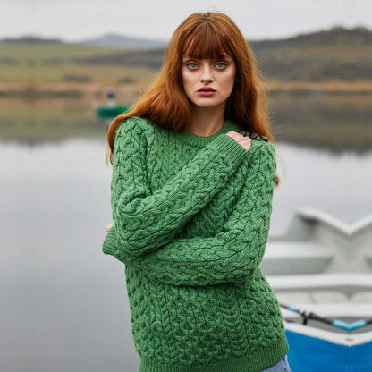 A woman with long red hair stands outdoors by a calm lake wearing  Ladies Side Button Aran Knit Sweater.  A boat is partially visible in the background, and the scene is softly blurred.