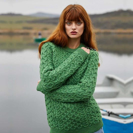 A woman with long red hair stands outdoors by a calm lake wearing  Ladies Side Button Aran Knit Sweater.  A boat is partially visible in the background, and the scene is softly blurred.
