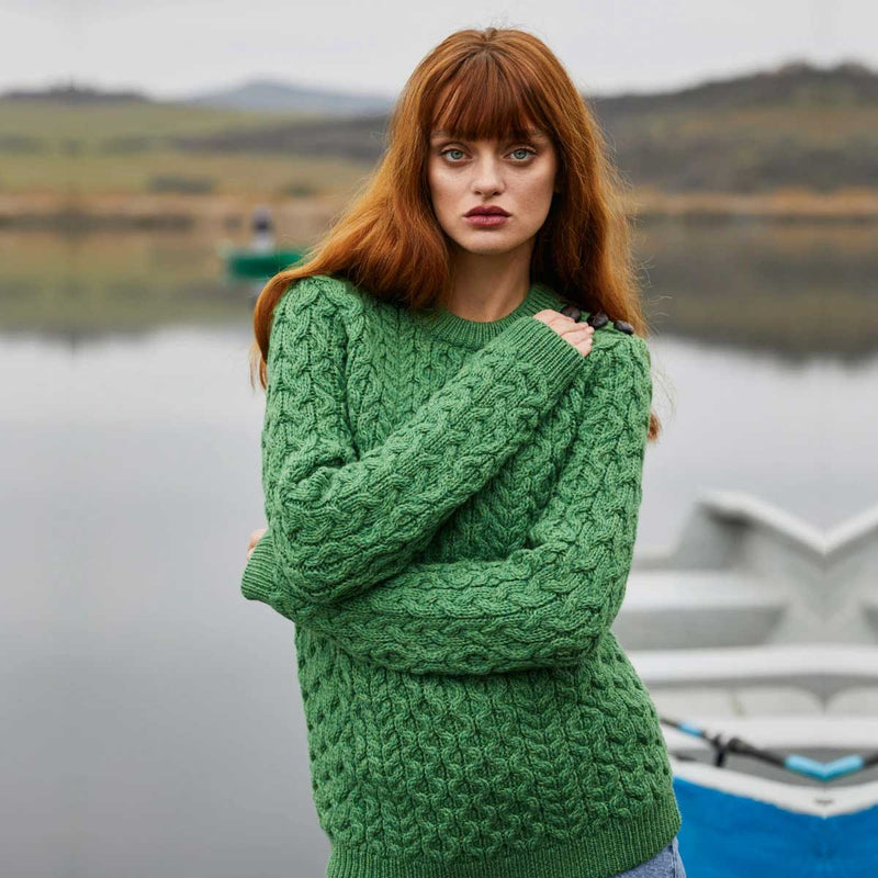 A woman with long red hair stands outdoors by a calm lake wearing  Ladies Side Button Aran Knit Sweater.  A boat is partially visible in the background, and the scene is softly blurred.