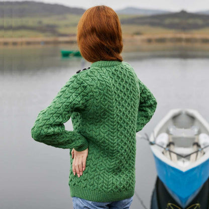 A person with red hair stands with their back to the camera wearing a  Ladies Side Button Aran Knit Sweater, standing next to a blue and white boat on a calm lake with hills in the background.