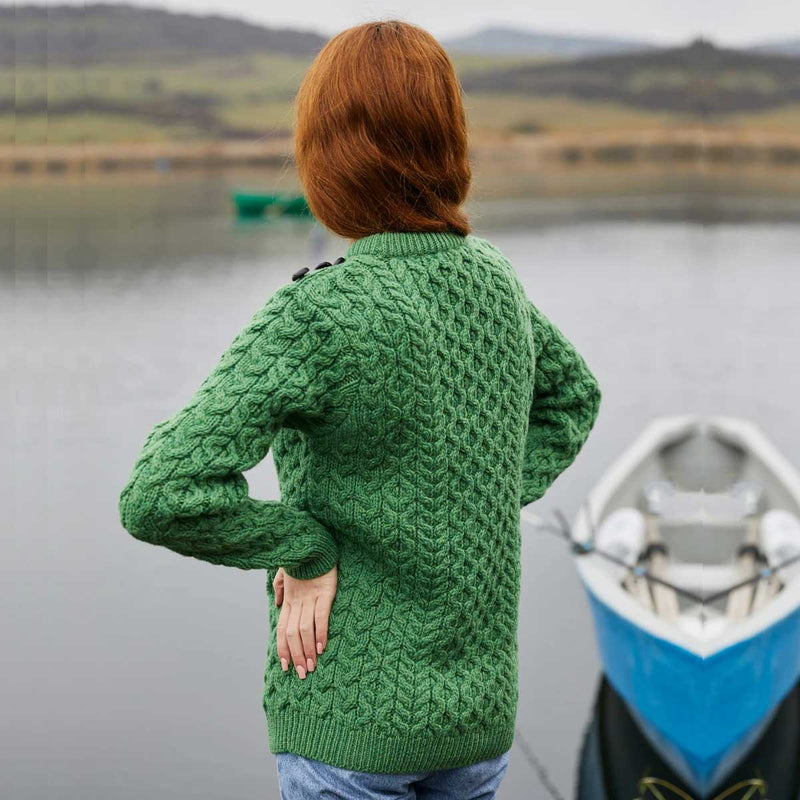 A person with red hair stands with their back to the camera wearing a  Ladies Side Button Aran Knit Sweater, standing next to a blue and white boat on a calm lake with hills in the background.